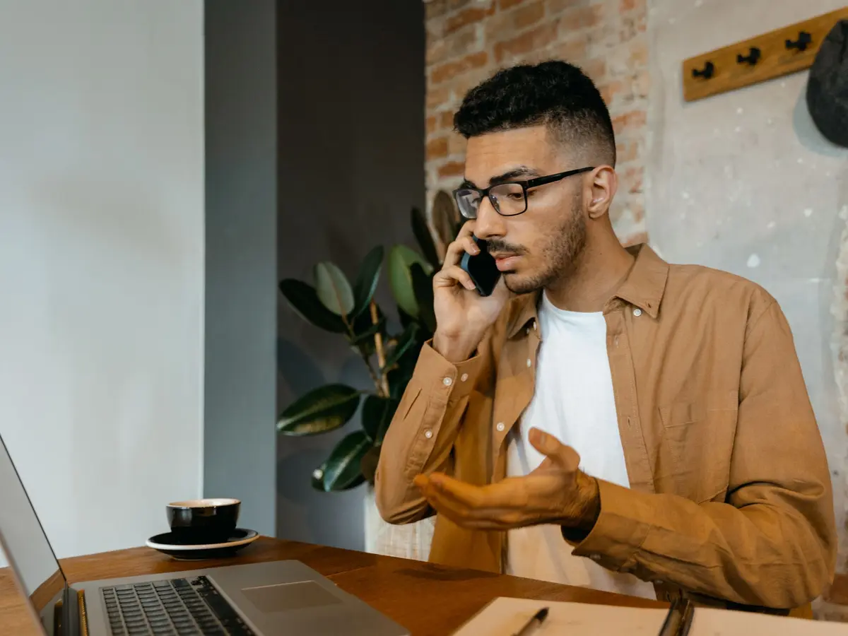 A man in glasses talks on the phone and gestures while working at a laptop on a wooden desk