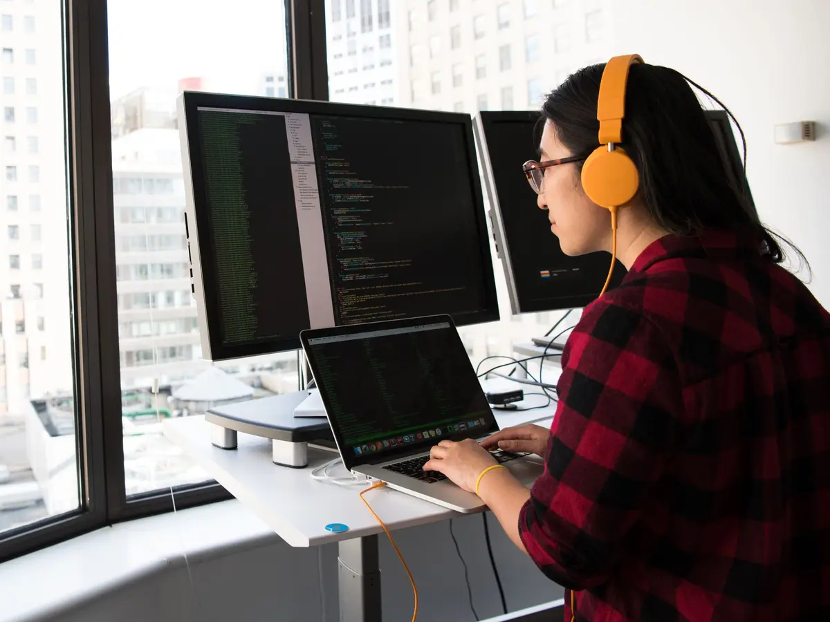 A developer wearing orange headphones writes code on a laptop and two monitors at a standing desk by a window