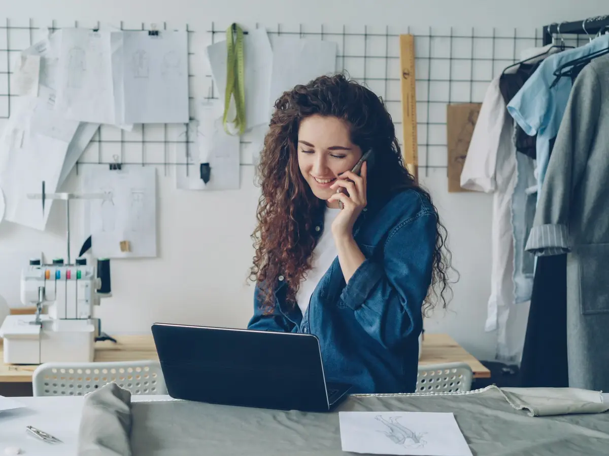 A fashion designer talks on her phone while working on a laptop in a bright sewing studio