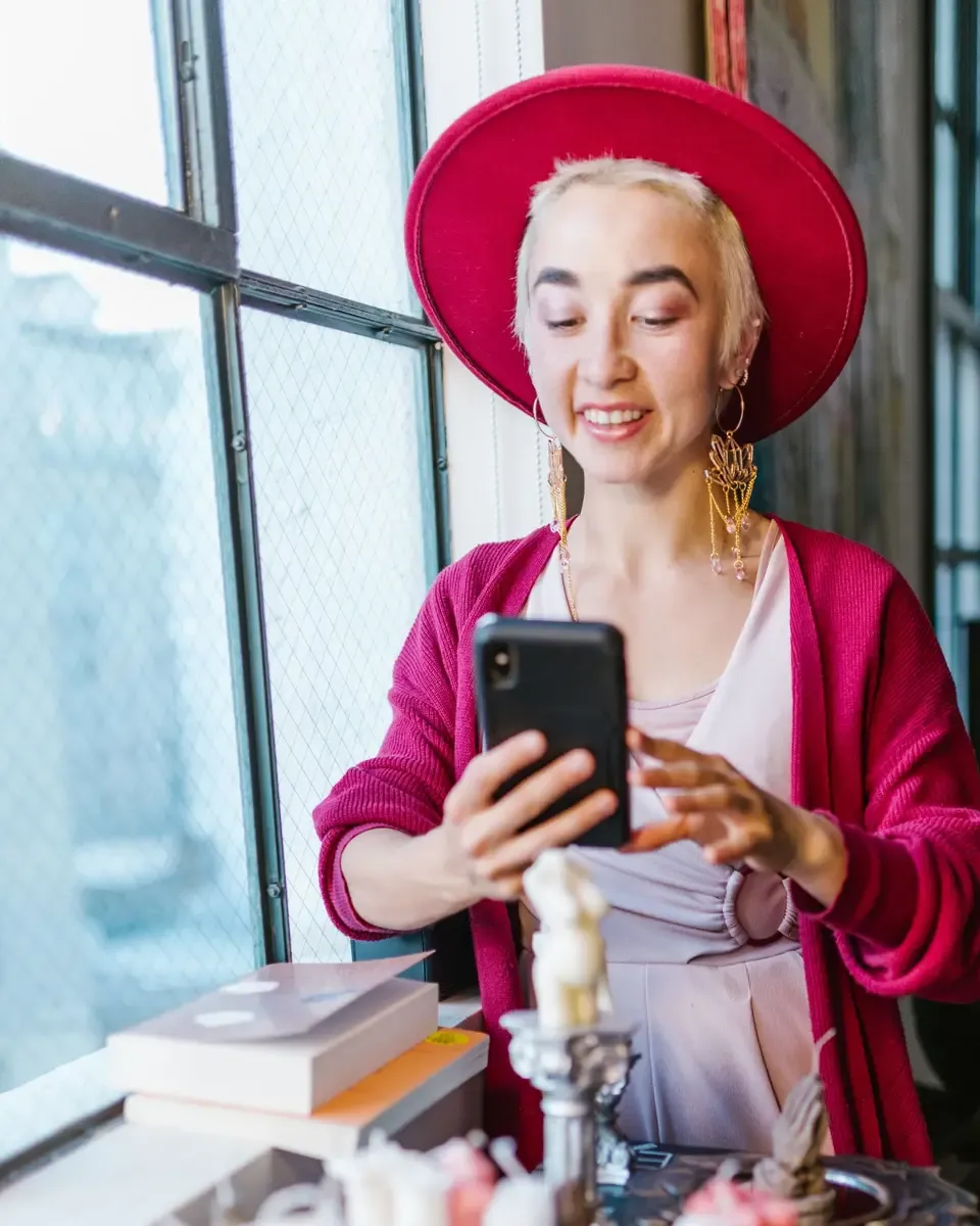 A woman in a red hat photographs her handmade candles with a phone by a loft window