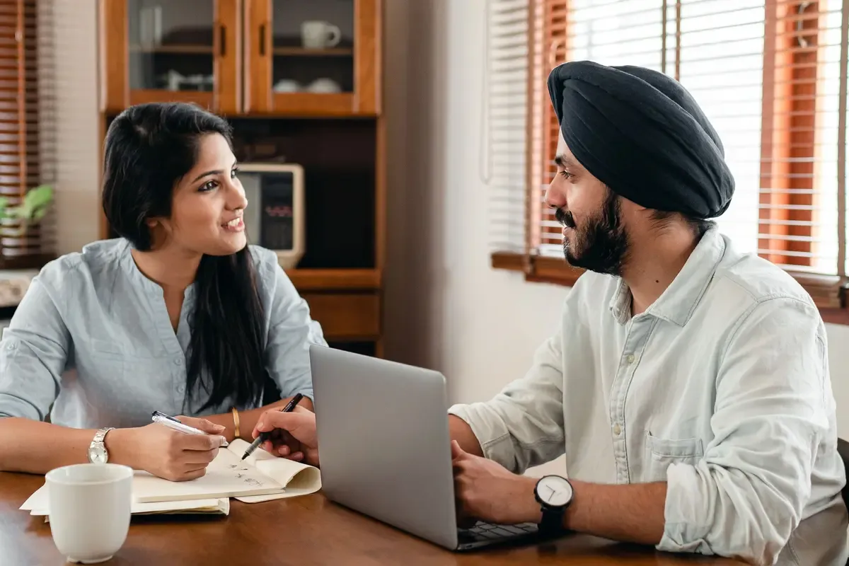 A woman taking notes and a man in a turban at a laptop smile at each other across a table