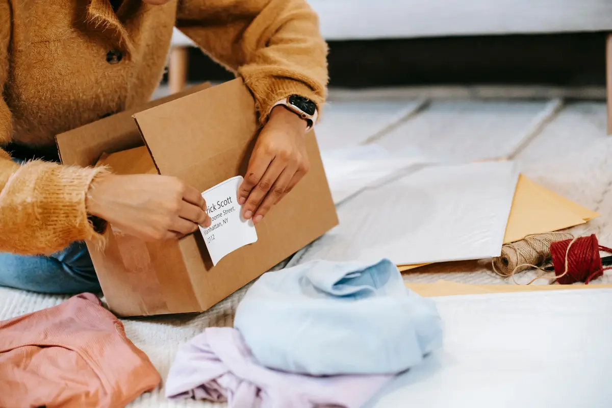 Hands stick an address label on a cardboard box beside folded clothes and mailers
