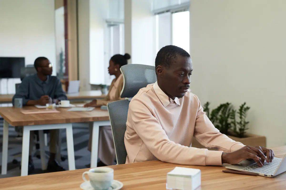 A man in a peach sweater works on a laptop at a long wooden desk in a bright open office