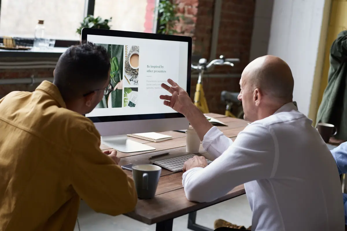 Two colleagues review a website layout on a desktop monitor, one pointing at the screen