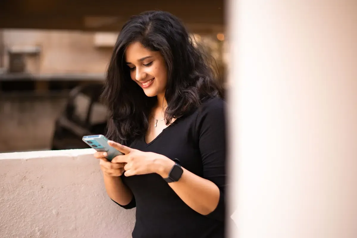 A young woman leans on a terrace wall, smiling as she types on her phone
