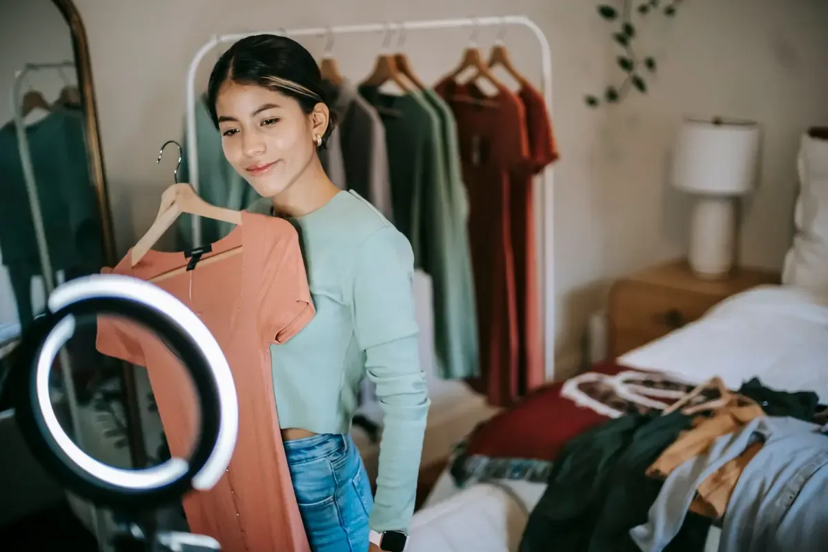 A young woman holds up a coral dress on a hanger in front of a ring light, clothing rail behind