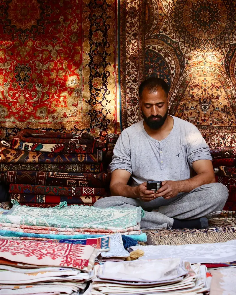A man sits cross-legged in his carpet and textile shop, typing on his phone