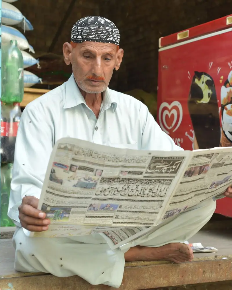 Shopkeeper in a white shalwar kameez and prayer cap reading an Urdu newspaper in his shop