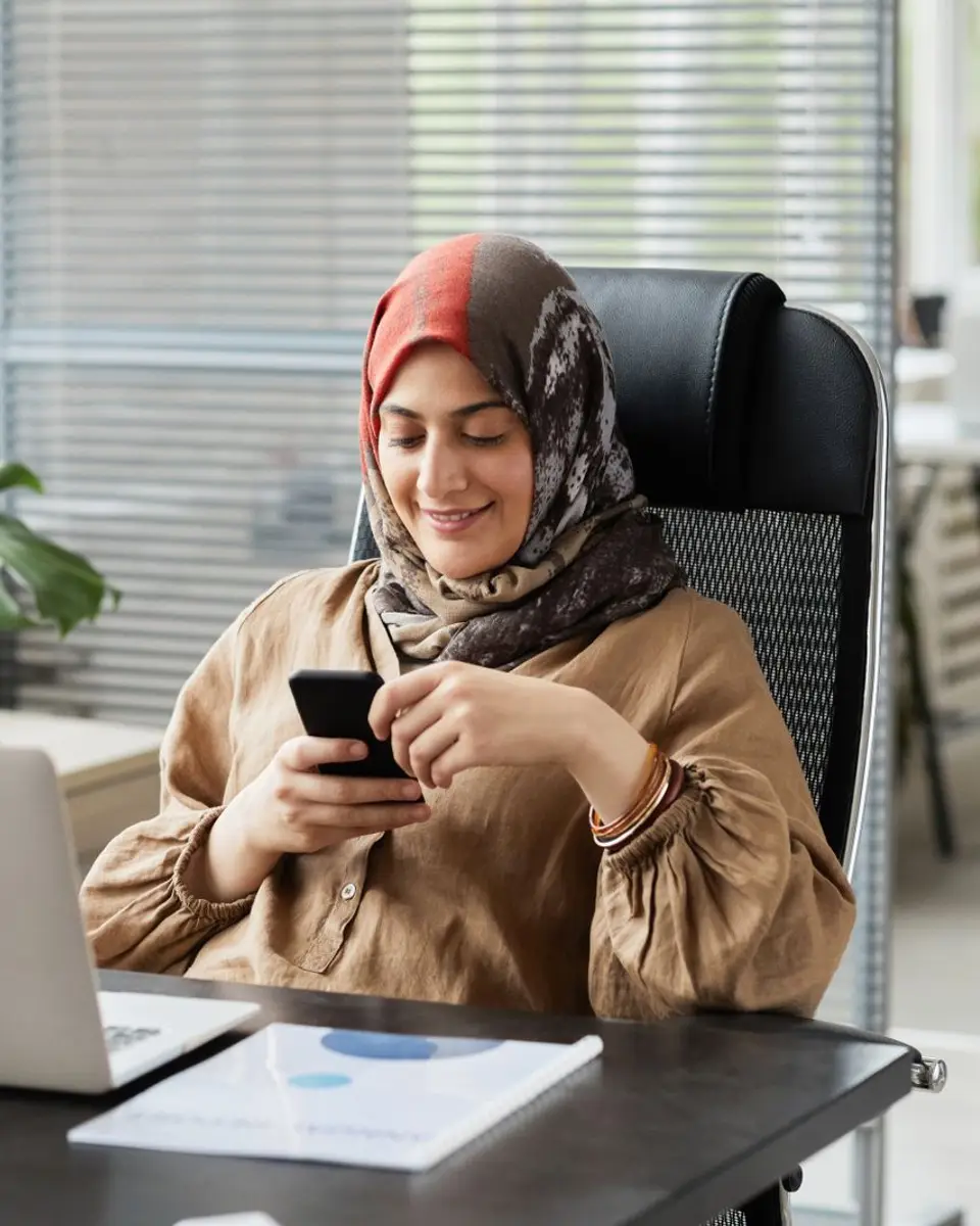 Business owner in a headscarf smiling at her phone beside her laptop in a bright office