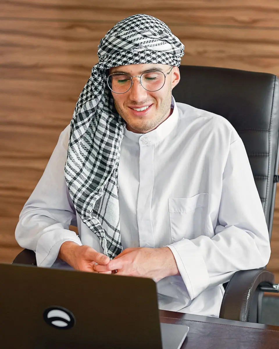 Smiling businessman in a white thobe and checked headscarf working on a laptop at his office desk