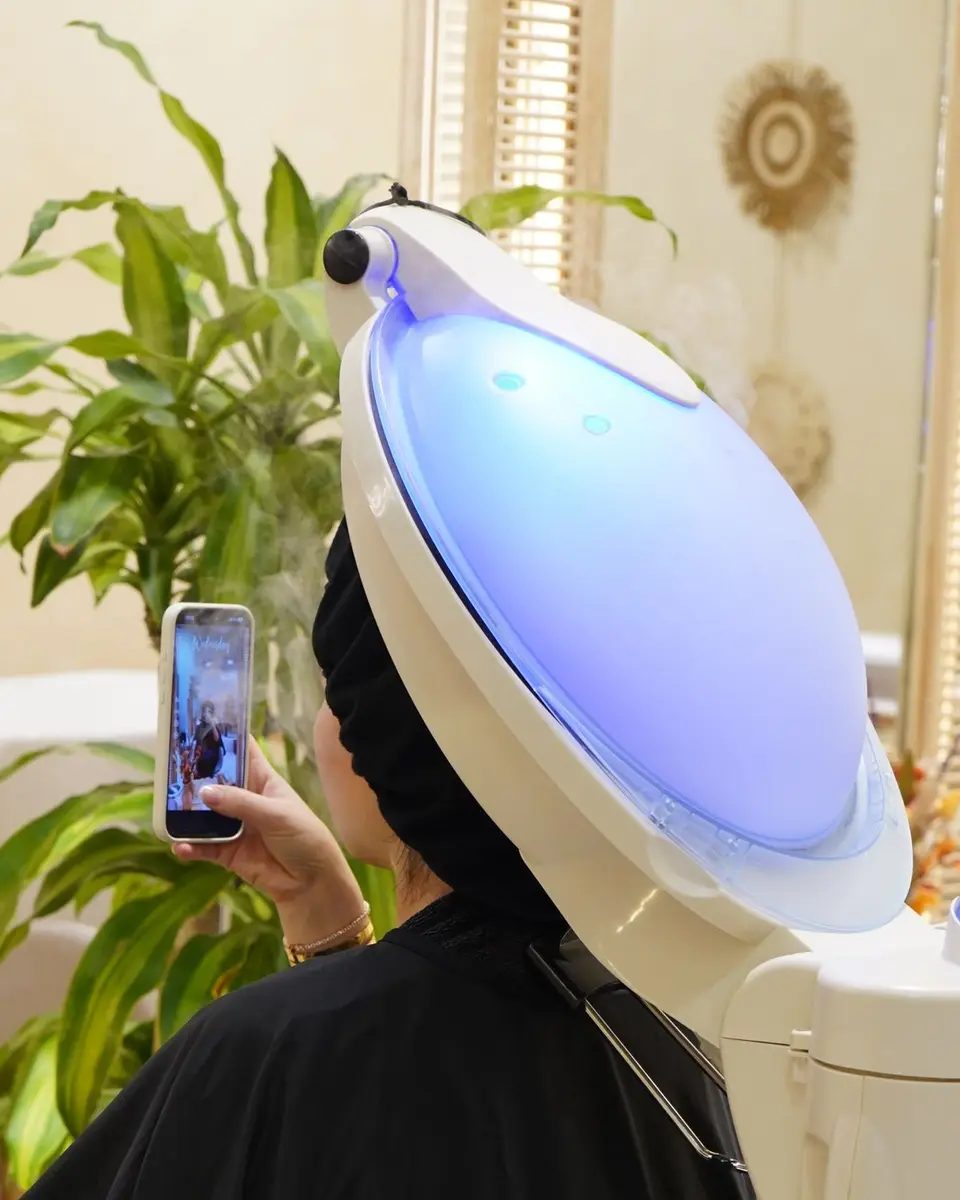 Salon client in a black headscarf looking at her phone while sitting under a hair steamer in a Dubai salon