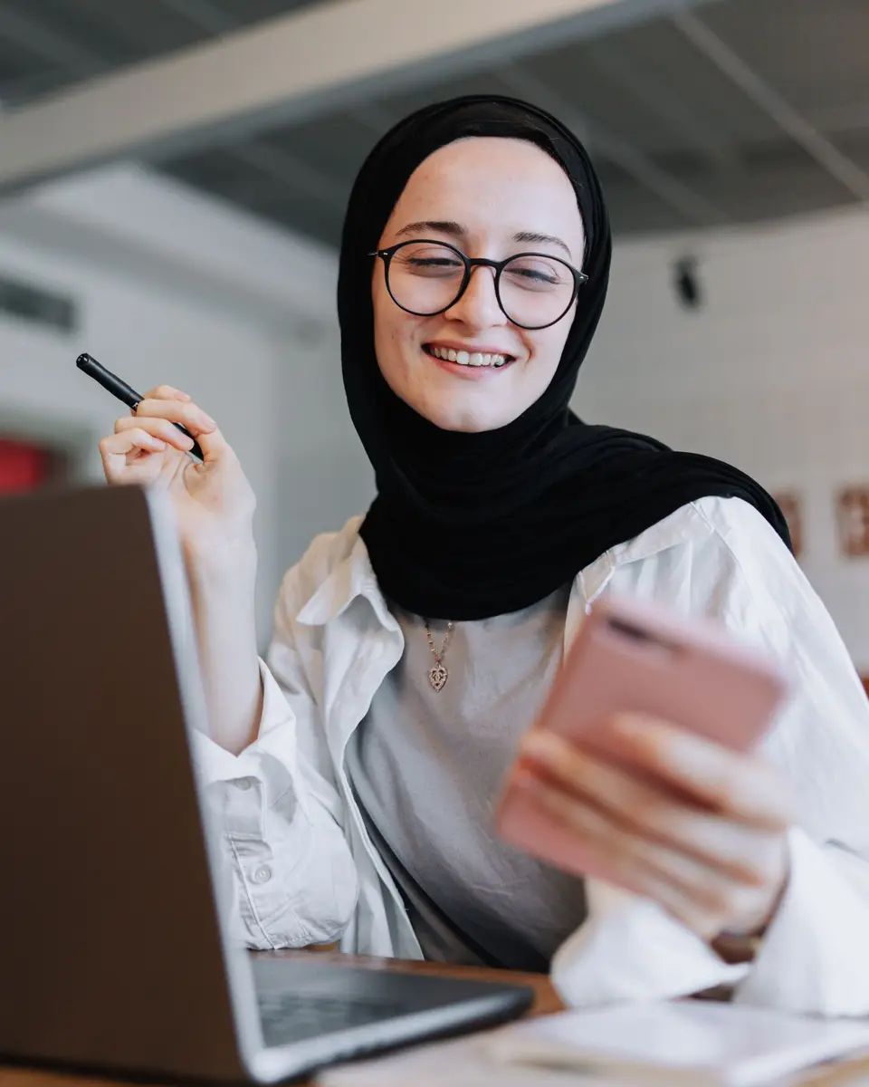 Smiling student in a black headscarf and glasses reading a reply on her phone beside her laptop