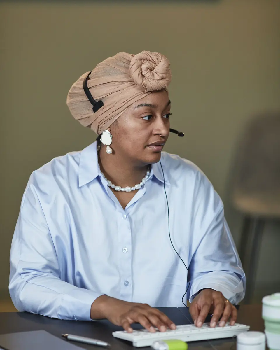 Support agent in a headset and a wrapped headscarf typing at her desk